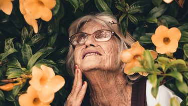 een oma met grijze haren voor een bosje met bloemen